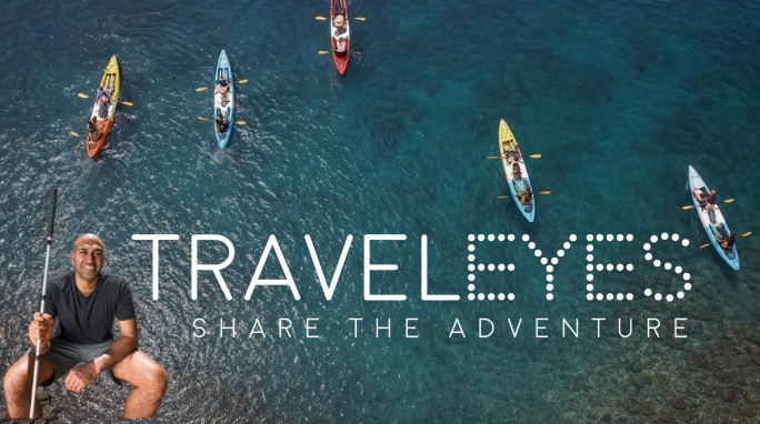 Photo of five 2-person kayaks in clear water with a photo of Amar Latif holding a white cane in one corner and the words 'TRAVELEYES SHARE THE ADVENTURE' in all capital letters in white across the mid-bottom of the photo.